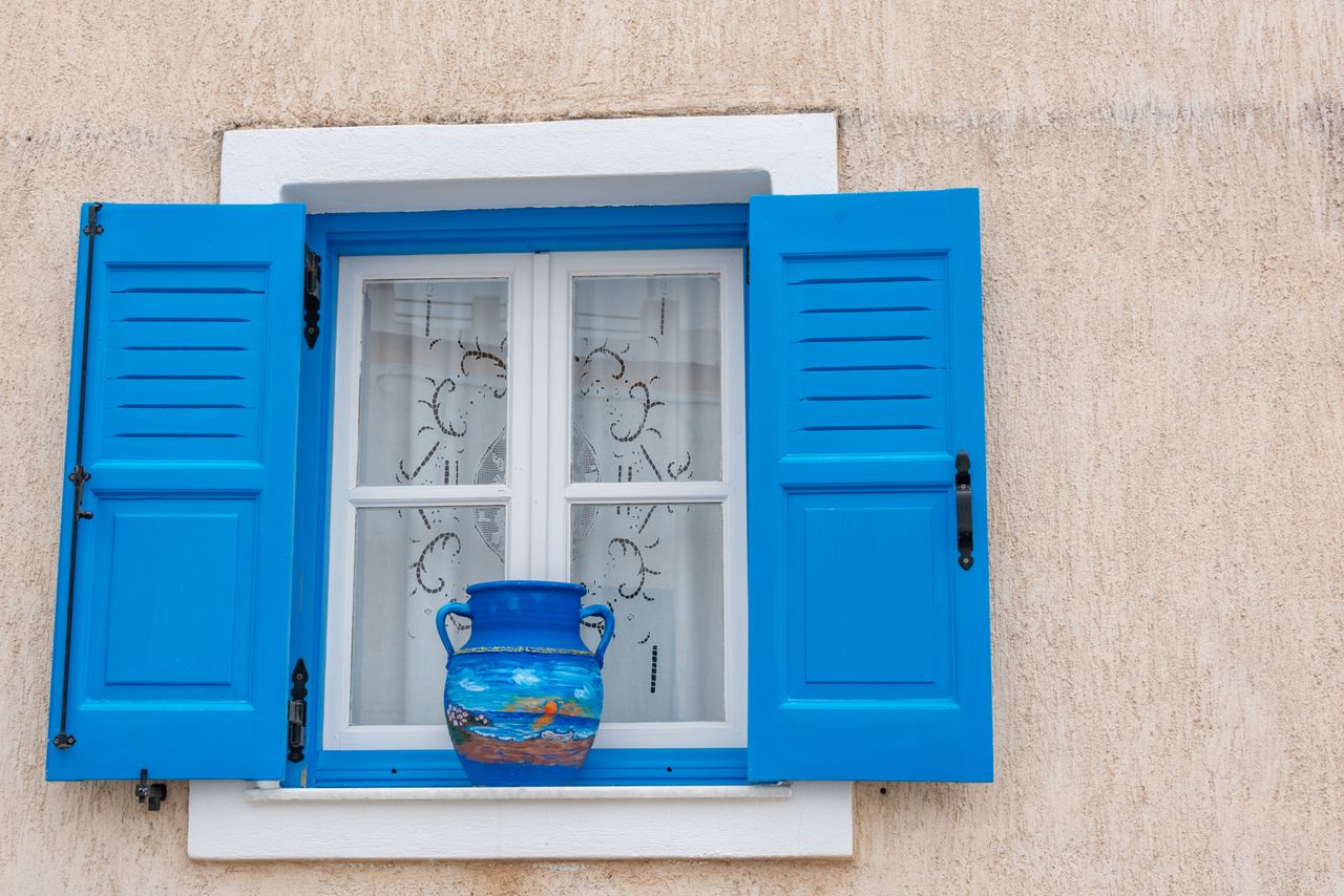 A vibrant painted vase on a windowsill framed by striking blue shutters, capturing a rustic charm.